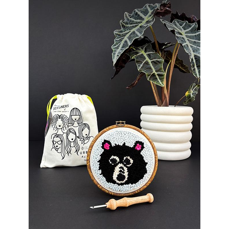 Embroidery hoop with a bear punch needle design, drawstring bag, and plant on a dark surface